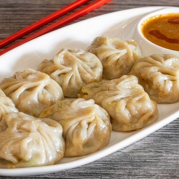 A white plate holds eight steamed momos with a side of orange dipping sauce and red chopsticks.