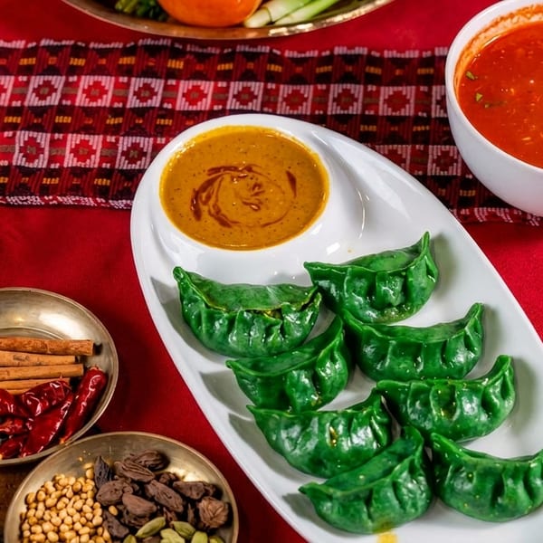 Green dumplings with dipping sauce, red soup, and bowls of spices on a red and patterned tablecloth.