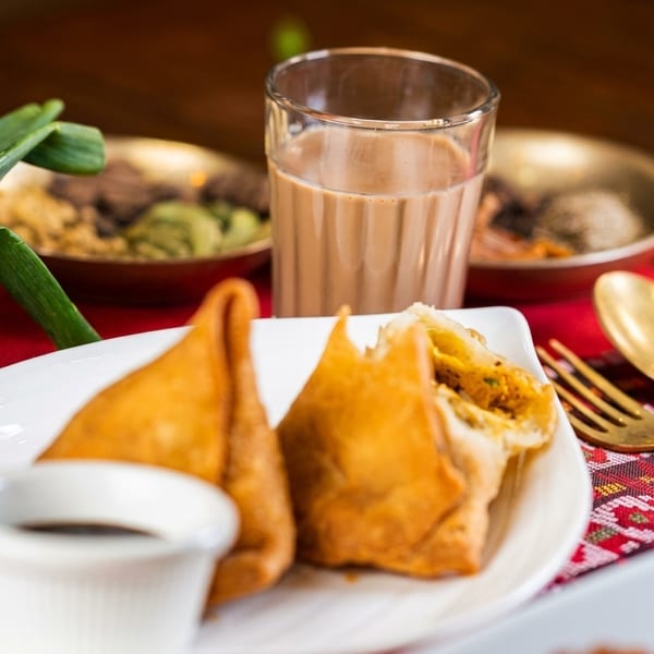 Two samosas, one broken open, with a dipping sauce, chai, and green onions on a red tablecloth.