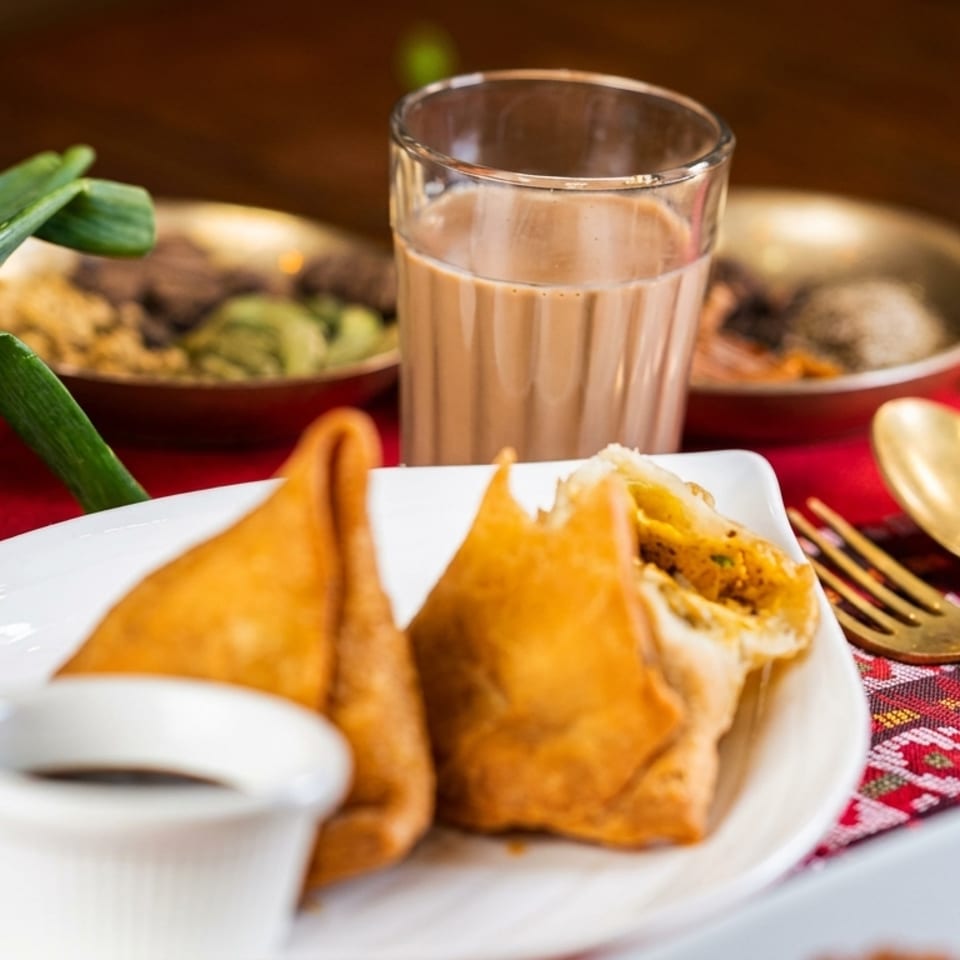 Two samosas on a white plate with dipping sauce, a glass of chai, and green onions.